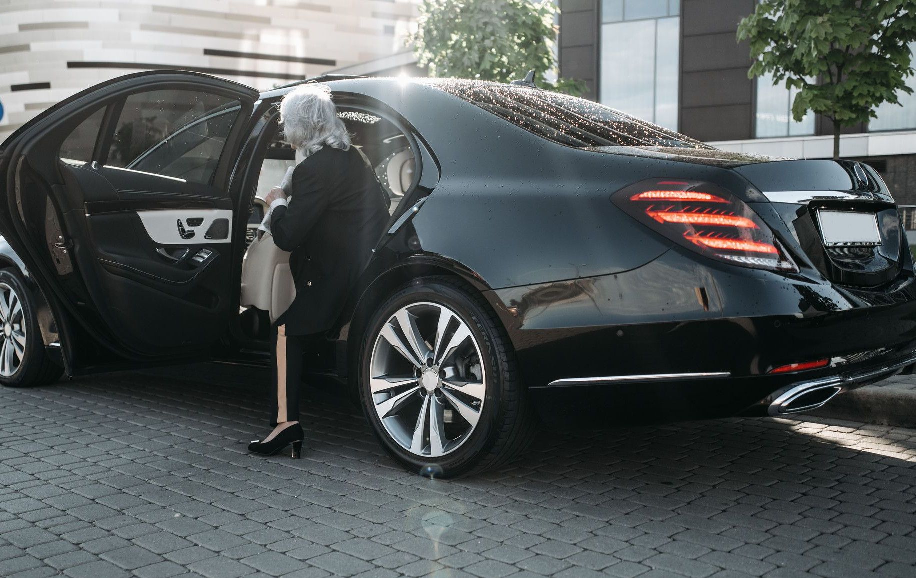 Senior businesswoman stepping out of a sleek black luxury vehicle in a cityscape.
